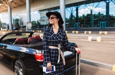 Beautiful smiling girl in sunglasses and black hat holding passport with flight ticket joyfully looking aside near airport with cabriolet car on background