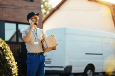 Young delivery man holding packages while communicating on mobile phone and standing on the street.